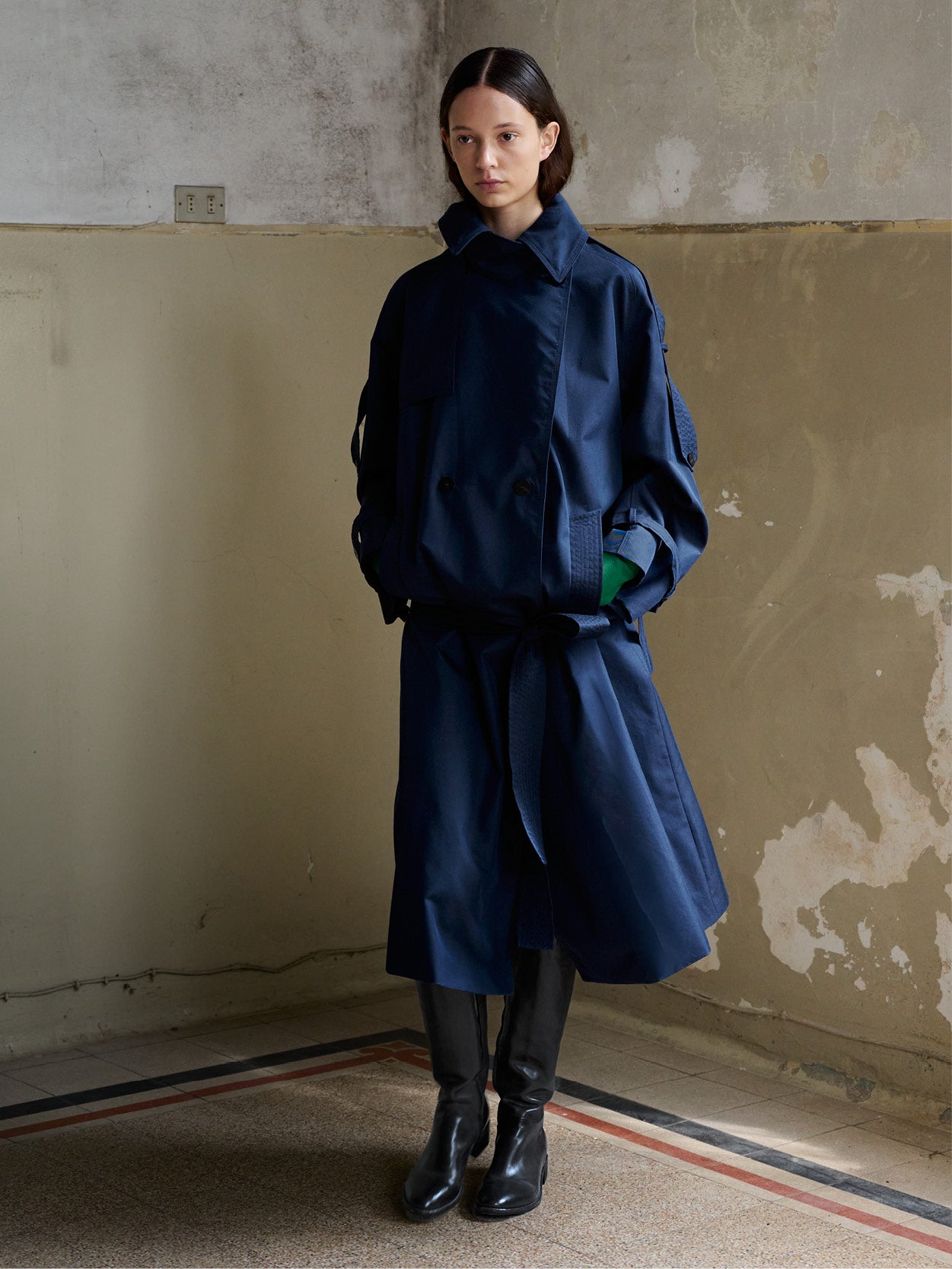 A model stands in a worn interior wearing a dark navy belted trench coat over a green top, paired with black boots.